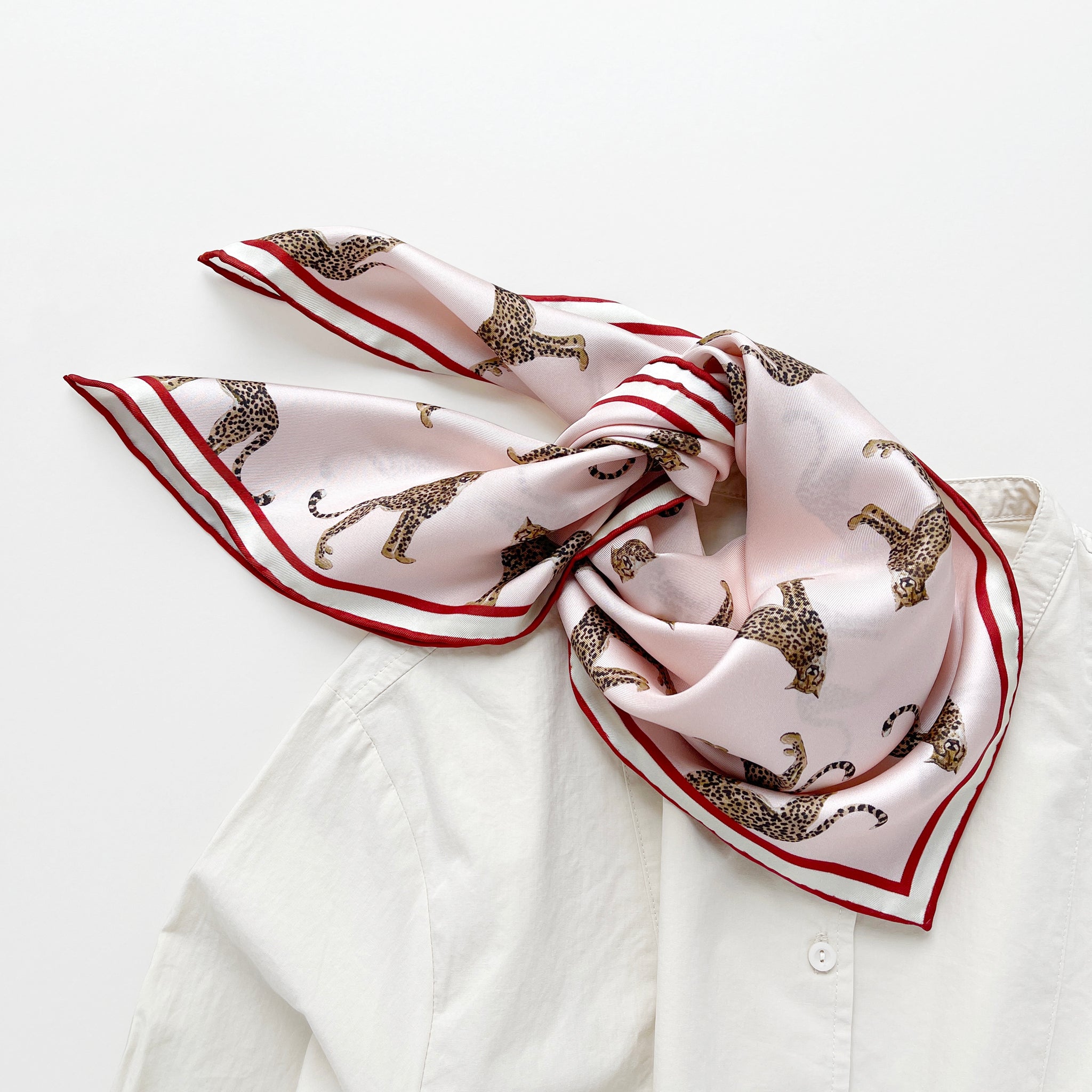 a pink base silk scarf featuring leopard print with red hand-rolled edges, knotted as a neck scarf, paired with a light beige women's shirt