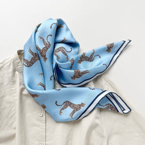 a sky blue base silk scarf featuring leopard print with dark blue hand-rolled edges, knotted as a neck scarf, paired with a light beige women's shirt