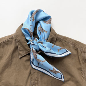 a sky blue base silk scarf featuring leopard print with dark blue hand-rolled edges, knotted as a neck scarf, paired with a khaki men's shirt