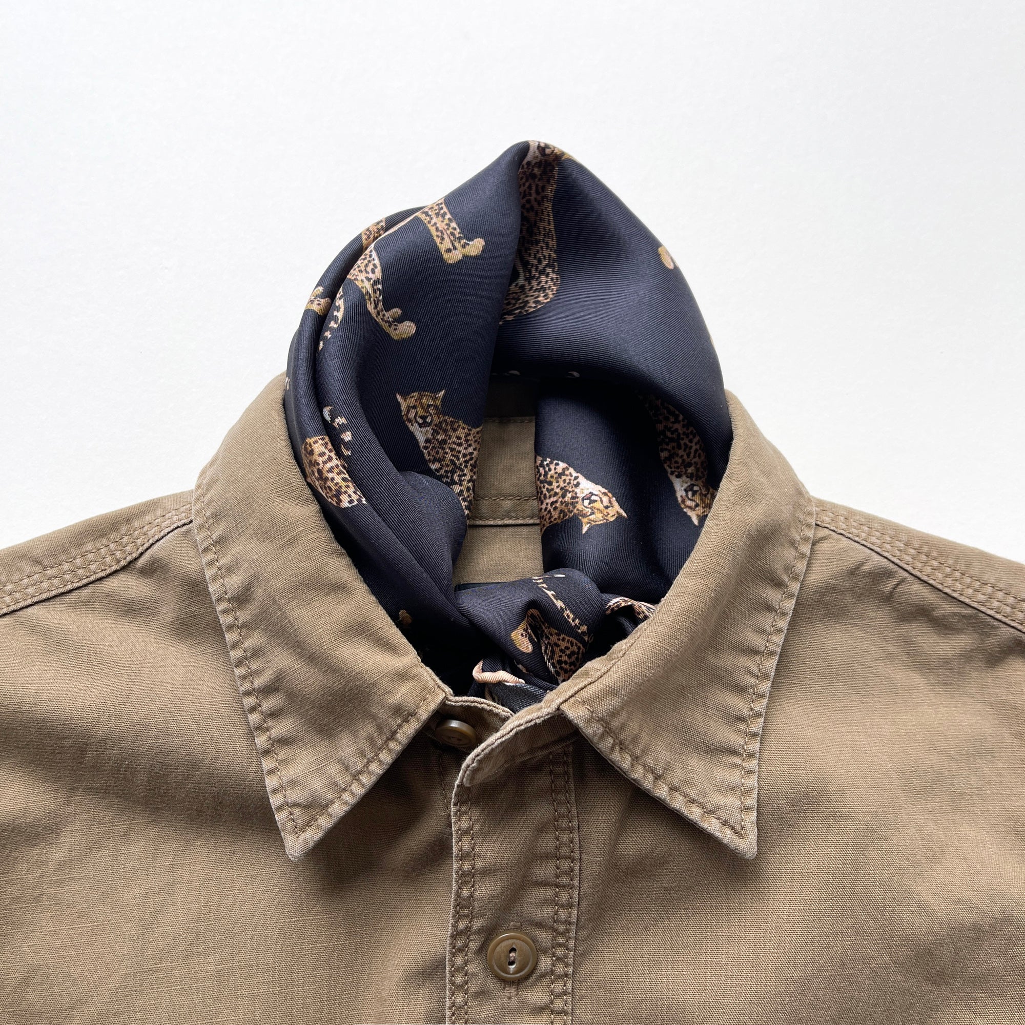 a black base silk scarf featuring leopard print with khaki brown hand-rolled edges, knotted as a neckerchief, paired with a khaki men's shirt 