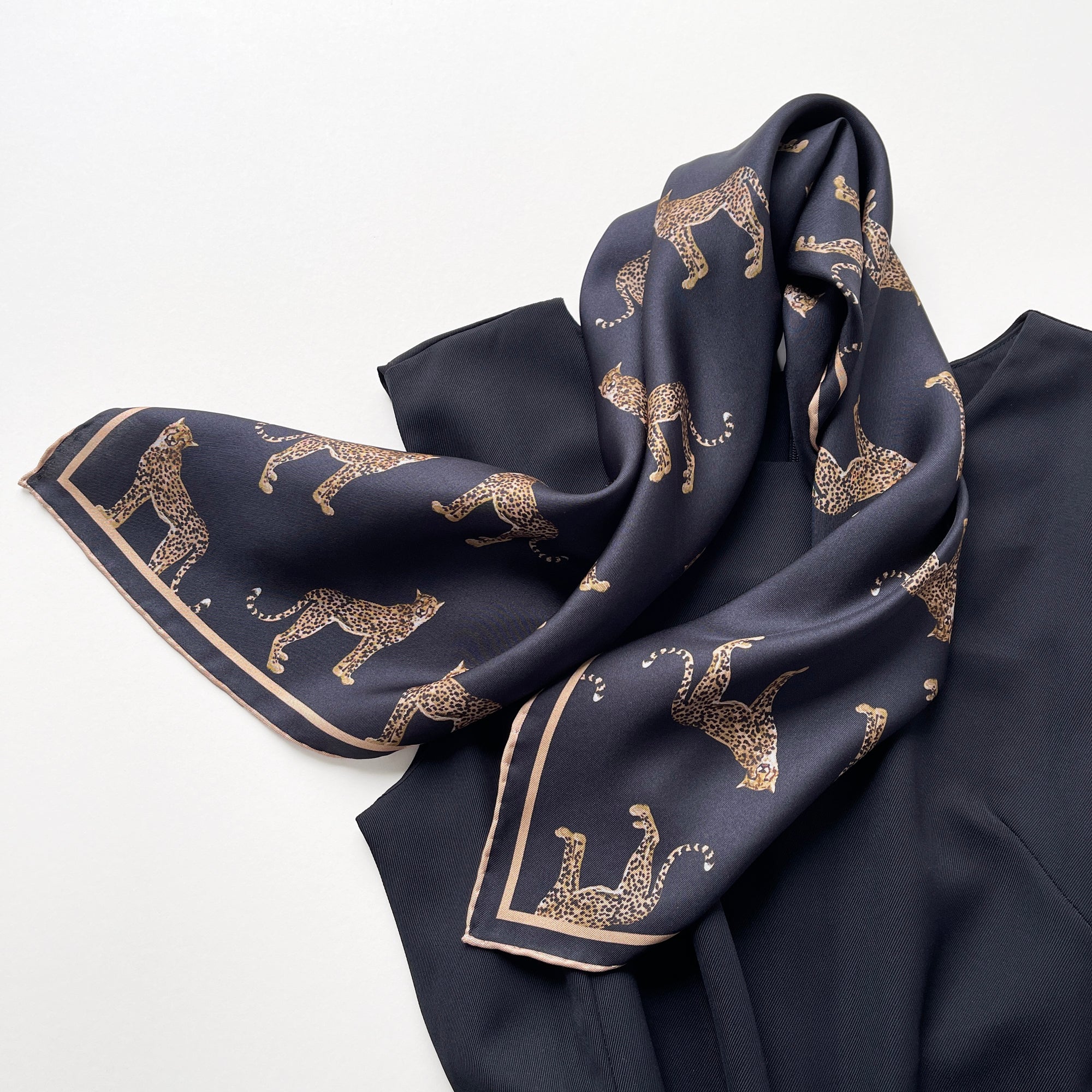 a black base silk scarf featuring leopard print with khaki brown hand-rolled edges, knotted as a neck scarf, paired with a women's black dress