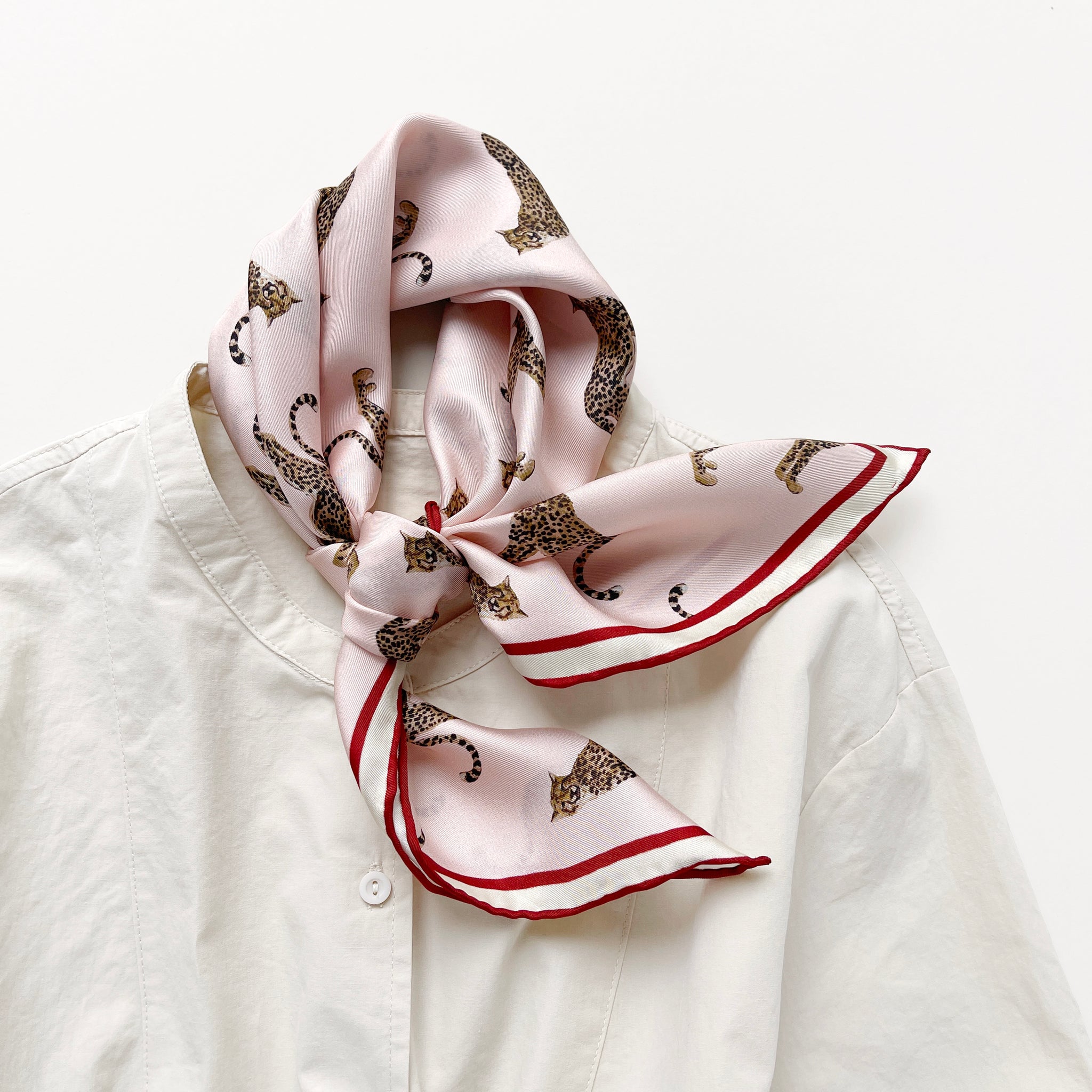 a pink base silk scarf featuring leopard print with red hand-rolled edges, knotted as a neck scarf, paired with a light beige women's shirt