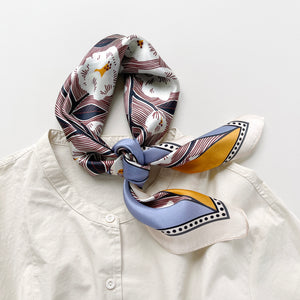 a women's silk scarf bandana featuring white rock roses print with cornflower blue and orange edges, knotted as a neck scarf, paired with a light beige turtleneck shirt