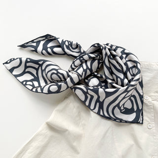 a white base women's silk scarf with deep charcoal grey abstract rose print and hand-rolled edges, knotted as a triangle bib, paired with a women's light beige turtleneck shirt