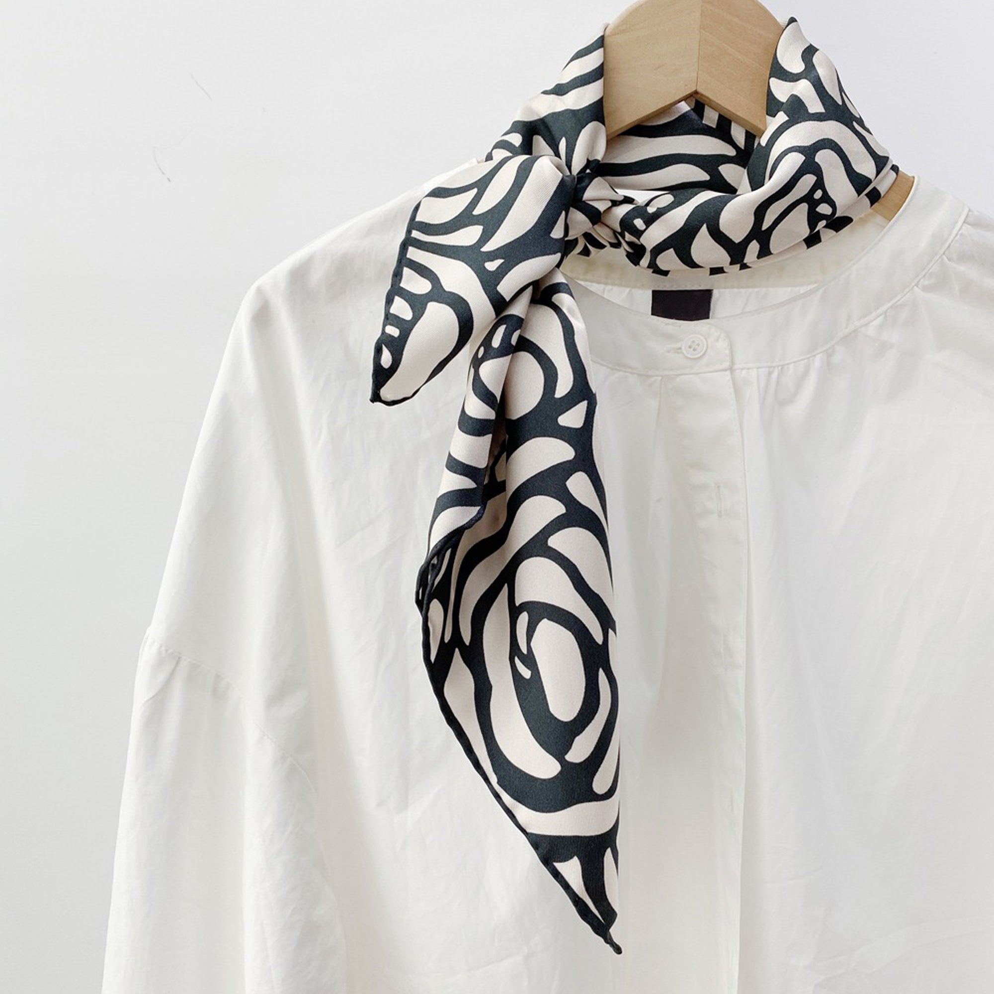 a white base women's silk scarf with deep charcoal grey abstract rose print and hand-rolled edges, knotted as a neck scarf, paired with a women's white turtleneck shirt 