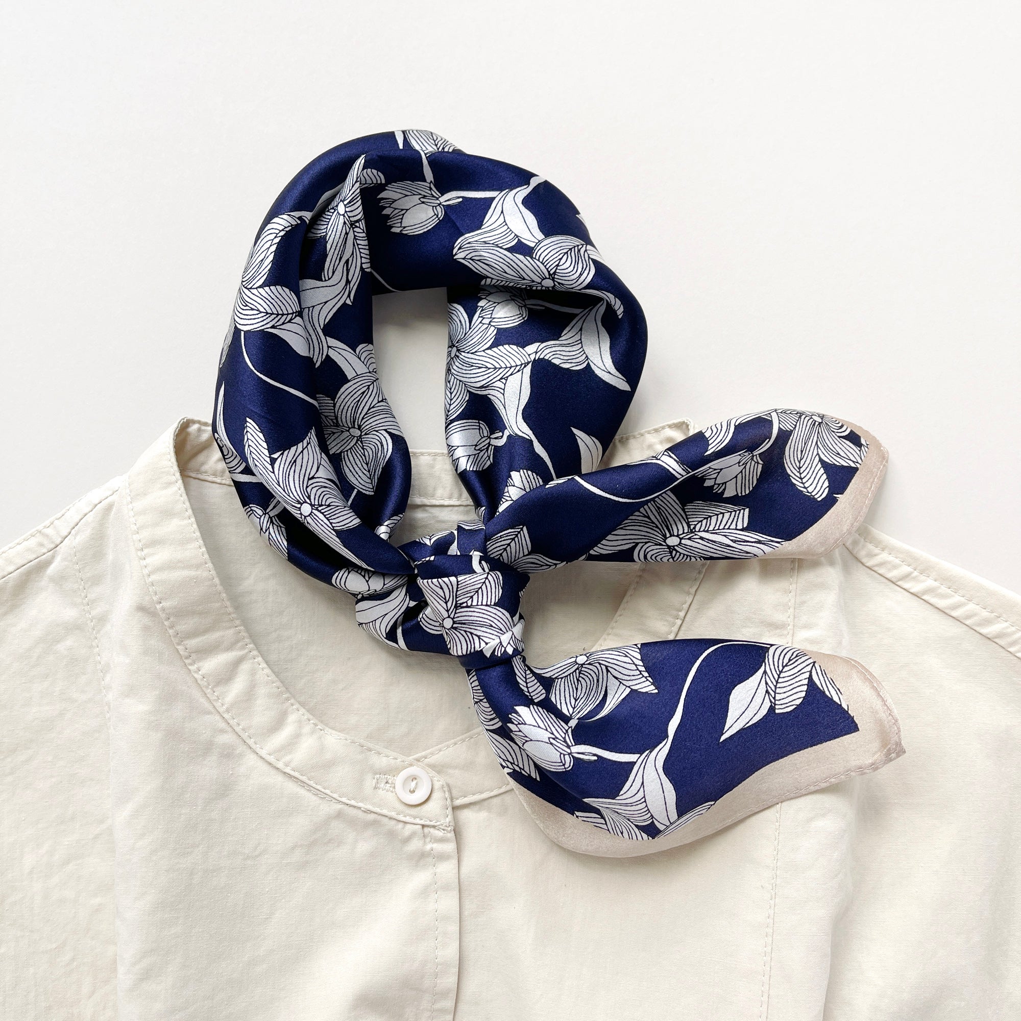 a deep blue silk bandana scarf featuring floral print with light beige edges, tied as a neck scarf, paired with a light beige turtleneck shirt