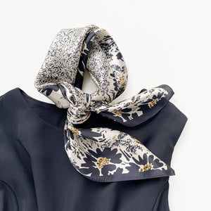 a women's black floral print silk scarf on light beige base, knotted as a neck scarf, paired with a black dress