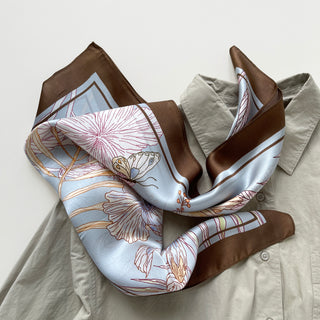 68x68 cm square silk scarf featuring a soft, light blue background with an intricate floral pattern of hibiscus flowers and butterflies. The flowers are outlined in shades of cream, yellow, and light brown, complemented by delicate leaves. The scarf has a wide, brown border framing the design.