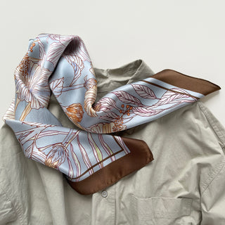 68x68 cm square silk scarf featuring a soft, light blue background with an intricate floral pattern of hibiscus flowers and butterflies. The flowers are outlined in shades of cream, yellow, and light brown, complemented by delicate leaves. The scarf has a wide, brown border framing the design.