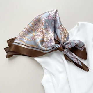 68x68 cm square silk scarf featuring a soft, light blue background with an intricate floral pattern of hibiscus flowers and butterflies. The flowers are outlined in shades of cream, yellow, and light brown, complemented by delicate leaves. The scarf has a wide, brown border framing the design.