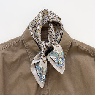 Men's silk neckerchief with a paisley and dotted pattern in beige, blue, and gold, styled around the neck of a khaki-colored shirt.