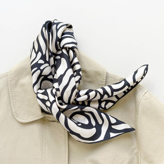 a white base women's silk scarf with deep charcoal grey abstract rose print and hand-rolled edges, knotted as a neck scarf, paired with a women's beige coat