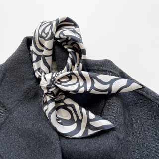 a white base women's silk scarf with deep charcoal grey abstract rose print and hand-rolled edges, knotted as a neck scarf, paired with a women's dark grey wool coat