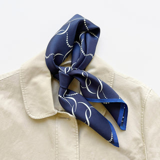 Navy blue silk scarf with a white geometric pattern, tied around the collar of a beige jacket. The 65x65 cm unisex scarf features hand-rolled edges and a smooth, luxurious finish, ideal for both men and women.