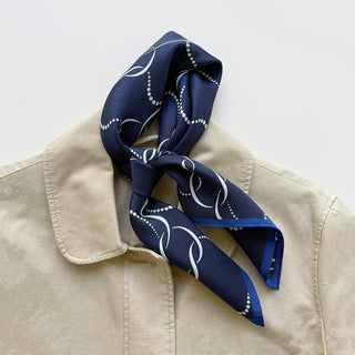 Navy blue silk scarf with a white geometric pattern, tied around the collar of a beige jacket. The 65x65 cm unisex scarf features hand-rolled edges and a smooth, luxurious finish, ideal for both men and women.