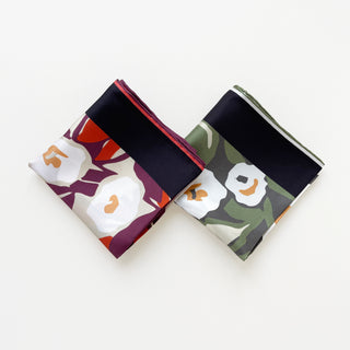 Two folded 100% silk twill bandanas with hand-rolled edges. One features a floral design with white flowers and orange leaves on a purple background, while the other has white flowers and green leaves on a darker background with a black border.