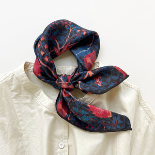 Deep blue silk bandana scarf with floral pattern tied around the neck. The scarf, made of 100% silk and featuring hand-rolled edges, displays red and pink floral designs, adding a chic touch to a white blouse. Ideal for styling "luxury silk neckerchief," "floral patterned scarf," and "elegant neck accessory."