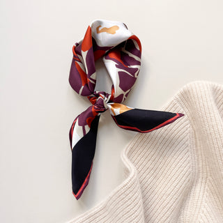 A silk twill bandana with a floral design in white, orange, and purple tones, tied in a knot and styled on a light beige cashmere sweater. The scarf's hand-rolled edges and vibrant colors are prominently displayed.