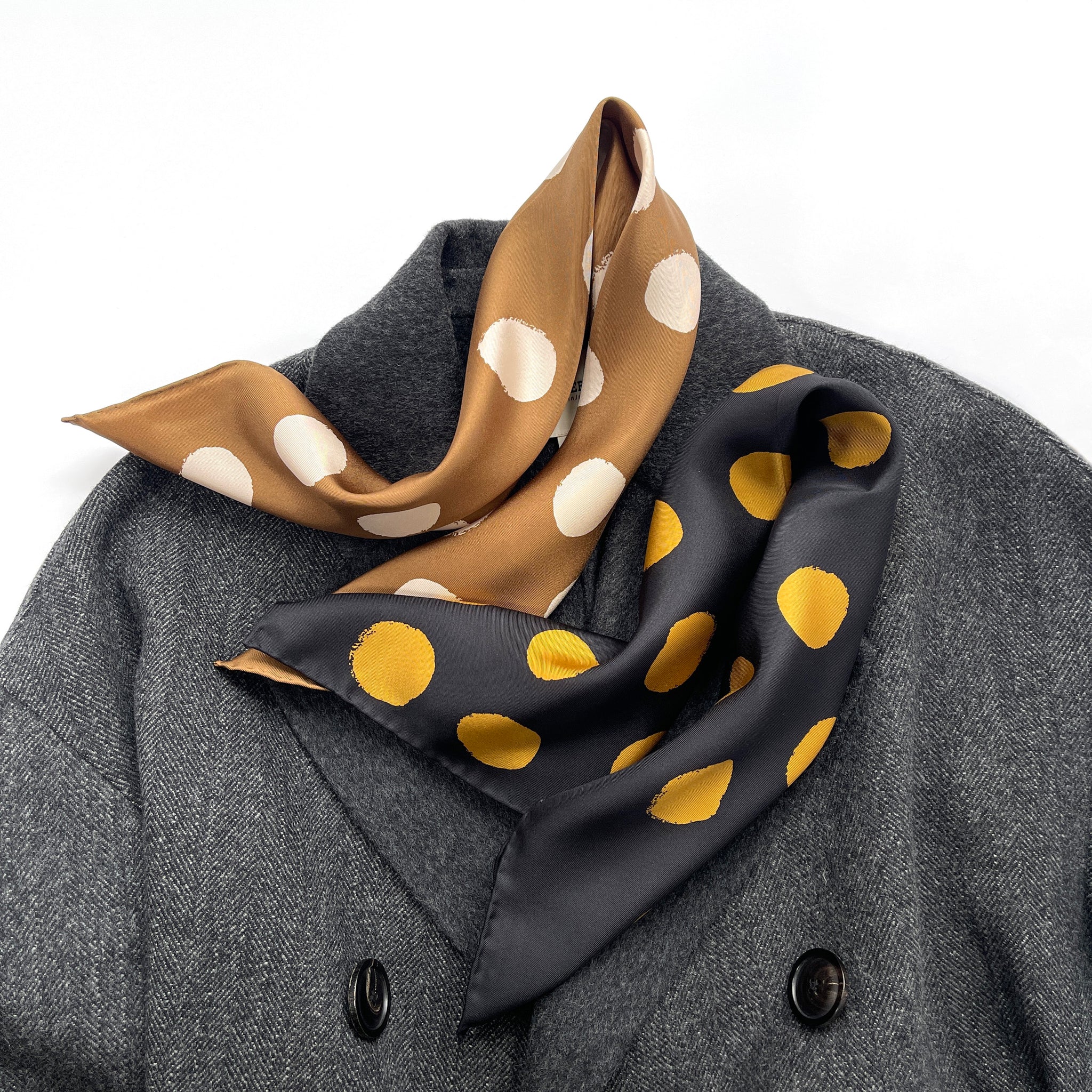 two polka dot print silk twill scarves in classic black and beige colours with hand-rolled hems laying on a dark grey wool coat