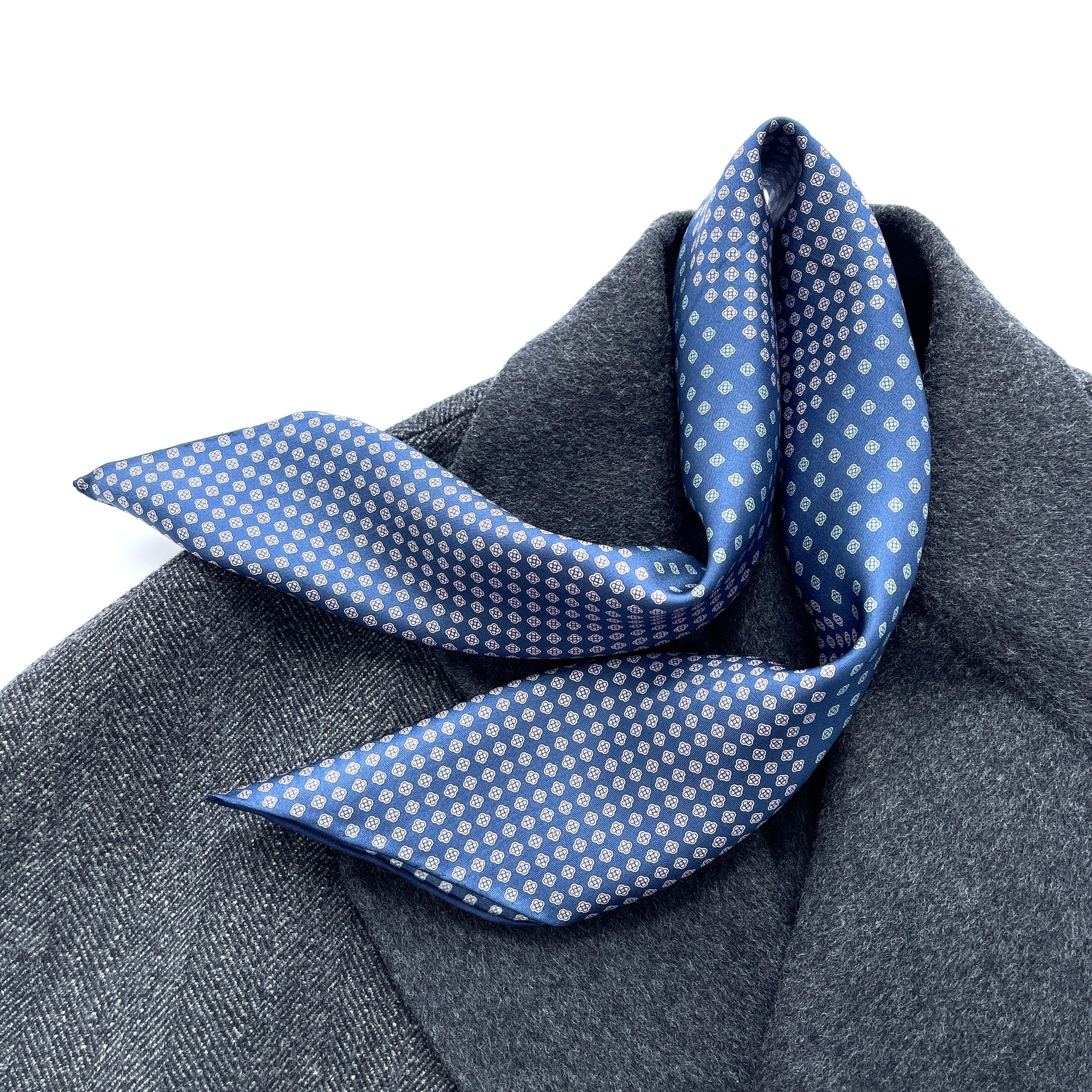 a navy blue men's silk scarf with hand-rolled hems featuring four leaf clover print knotted as a neckerchief, laying on a dark grey wool coat