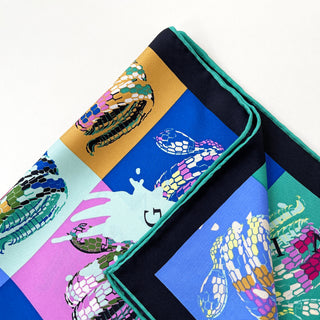 A vibrant folded square silk scarf with swirling snakes print and hand-rolled hems. The swirling snakes are printed on different colour blocks such as blue, red, green, and yellow.