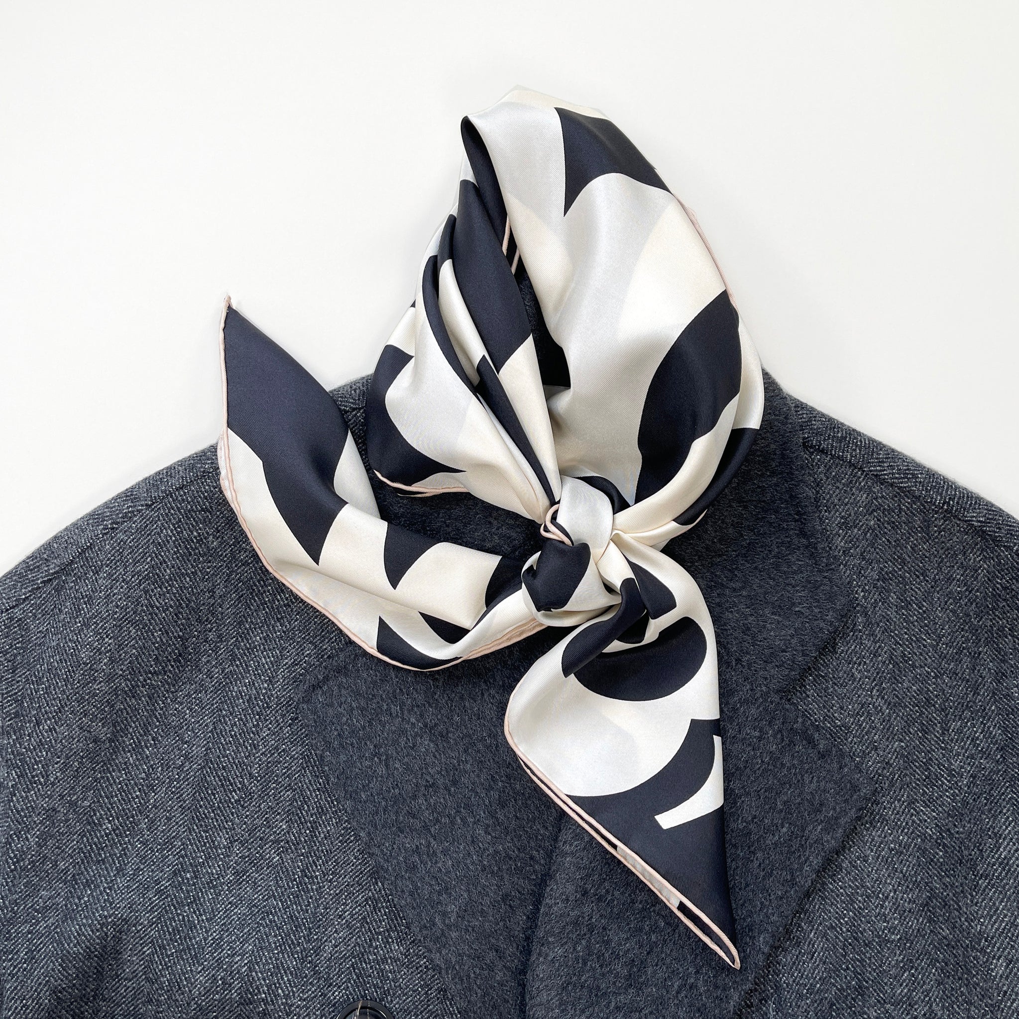 a black and white large square silk scarf with camellia print knotted as a neck scarf laying on a dark grey wool coat