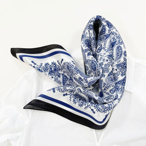 a white based silk scarf with rich blue paisley pattern print featuring black edge laying on a white shirt