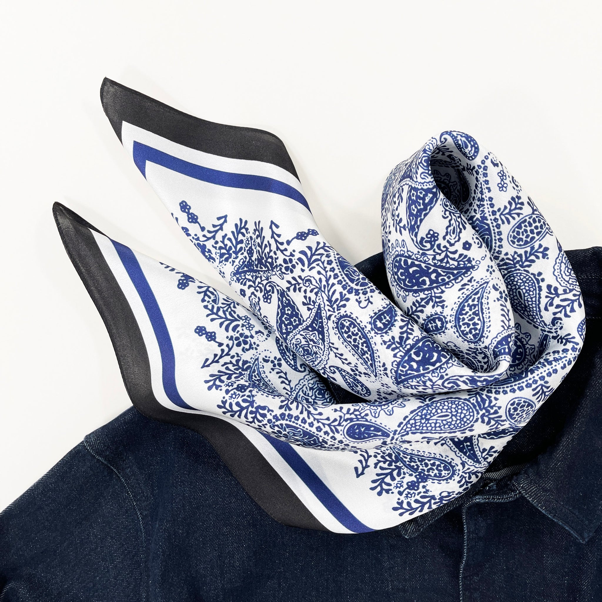 a white based silk scarf with rich blue paisley pattern print featuring black edge laying on a deep blue men's denim jacket