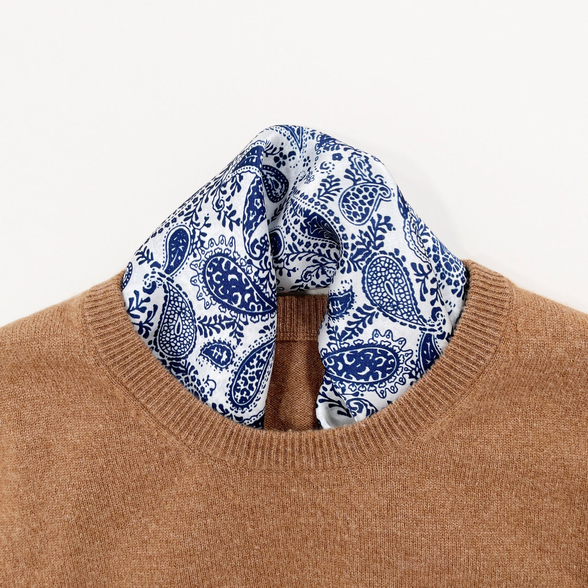 a white based silk scarf with rich blue paisley pattern print featuring black edge tucked into a camel men's sweater