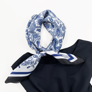 a white based silk scarf with rich blue paisley pattern print featuring black edge knotted as a neck scarf laying on a woman's sleeveless black dress