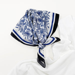 a white based silk scarf with rich blue paisley pattern print featuring black edge with a white sleeveless woman's dress