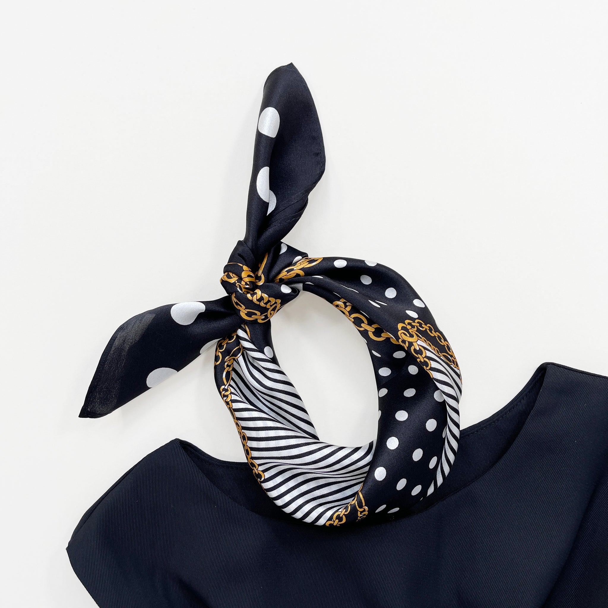 a small square silk scarf for women featuring polka dot and stripe print in black and white palette knotted as a headscarf paired with a sleeveless black dress