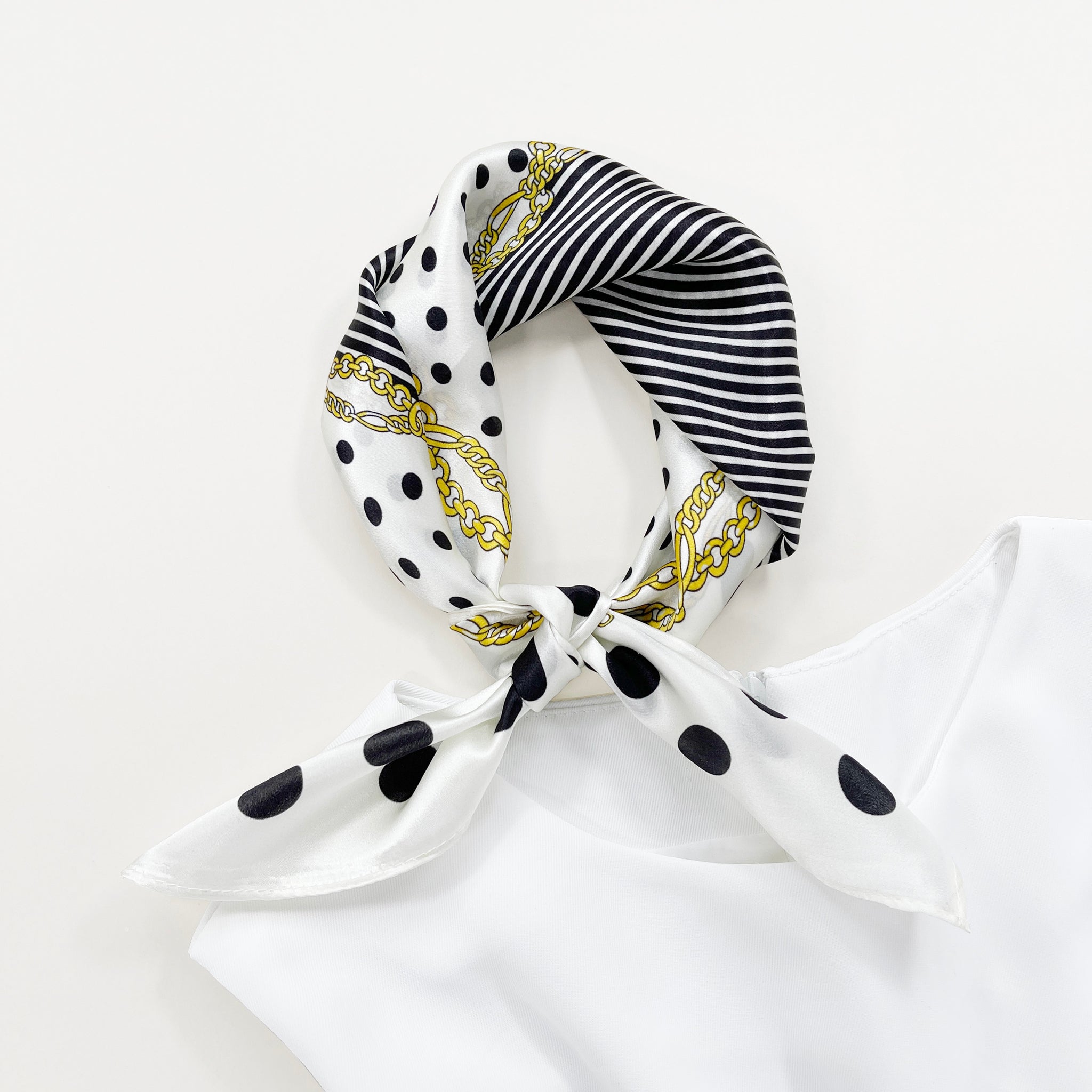 a small square silk scarf for women featuring polka dot and stripe print in white and black palette knotted as a neckerchief paired with a sleeveless white dress