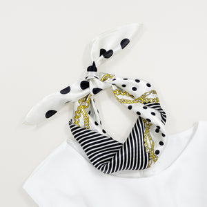 a small square silk scarf for women featuring polka dot and stripe print in white and black palette knotted as a neck scarf or headscarf paired with a sleeveless white dress