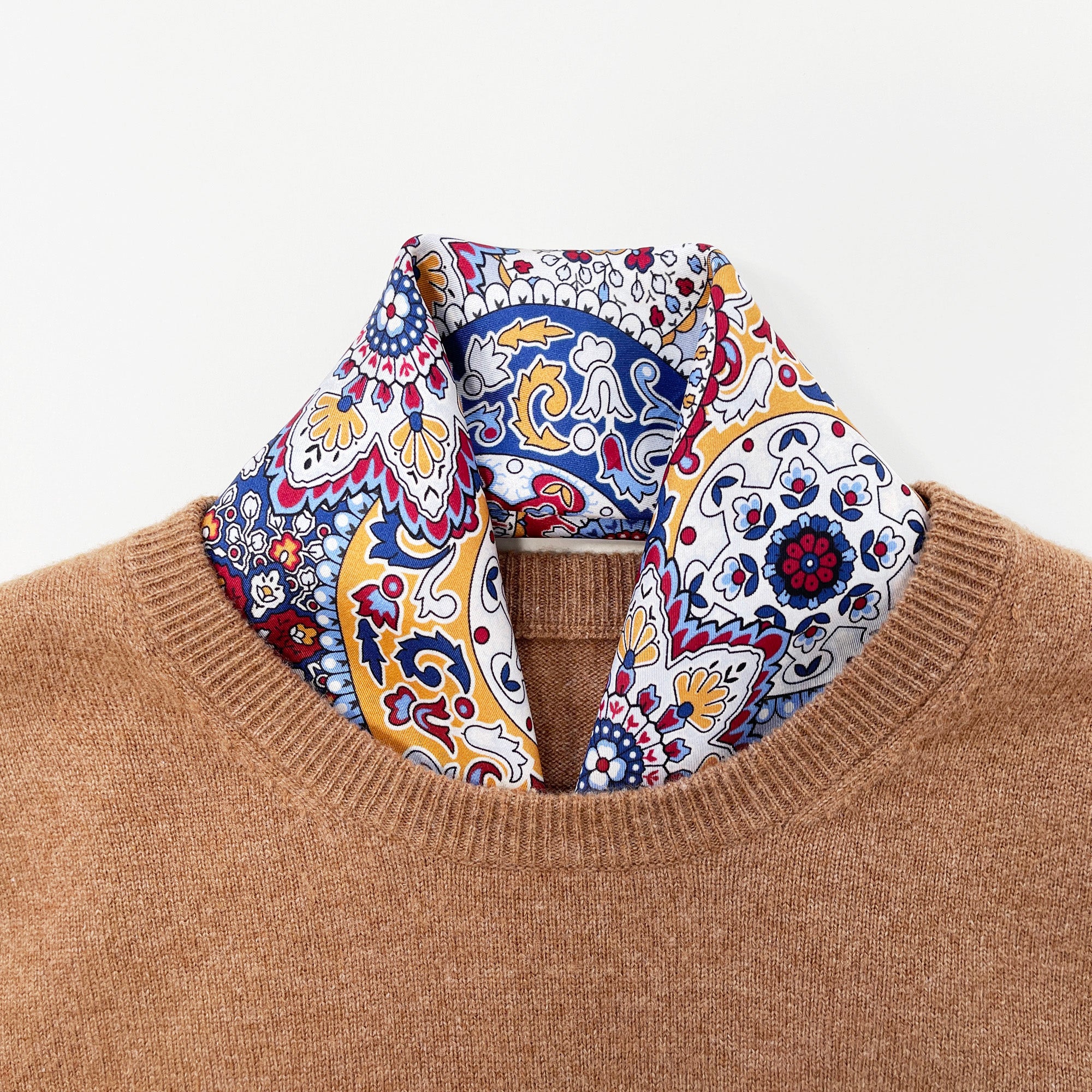 a red, blue and yellow silk scarf square for men featuring boho-style print and red hand-rolled hems tucked in a camel sweater