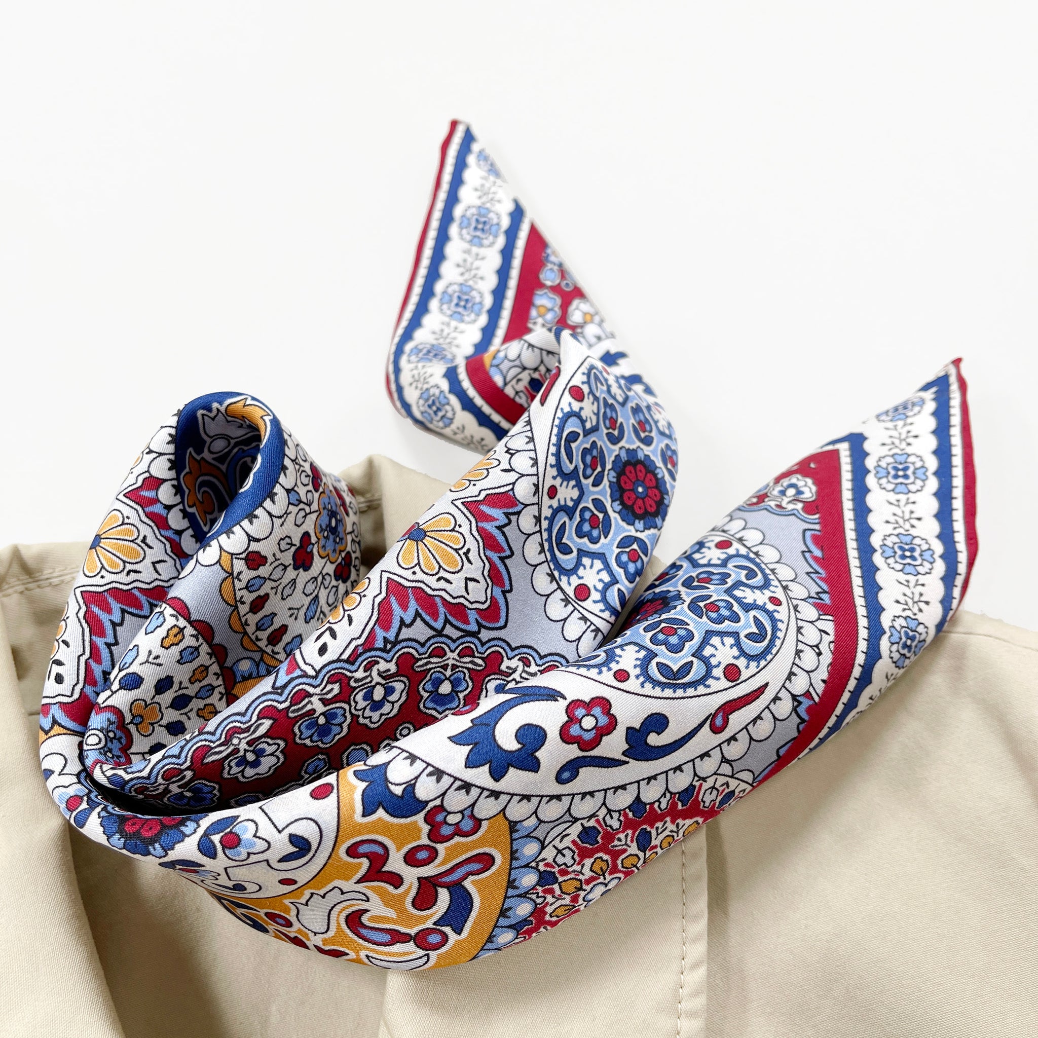 a red, blue and yellow silk scarf square for men featuring boho-style print and red hand-rolled hems laying on a beige coat