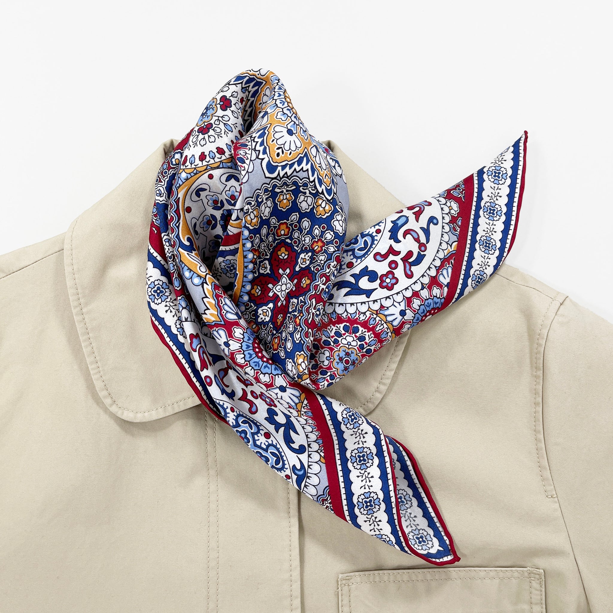a red, blue and yellow silk scarf square for men featuring boho-style print and red hand-rolled hems laying on a beige coat