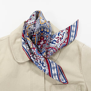 a red, blue and yellow silk scarf square for men featuring boho-style print and red hand-rolled hems laying on a beige coat