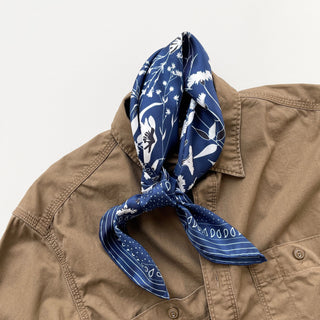 a rich blue men's silk scarf featuring botanic leafy print and hand-rolled hems knotted as a neckerchief, paired with a khaki shirt