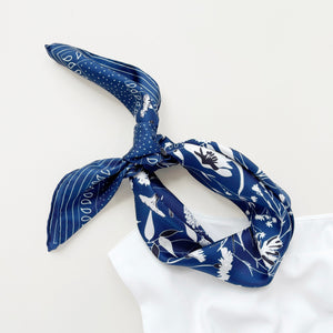 a rich blue silk scarf featuring botanic leafy print and hand-rolled hems knotted as a headband, paired with a white sleeveless dress