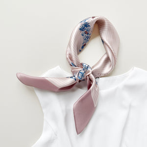 a blush pink silk scarf for women featuring blue and burgundy leafy print knotted as a neck scarf, paired with a white sleeveless dress