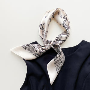 a light beige silk scarf featuring black paisley print knotted as a necks scarf, paired with a black sleeveless dress