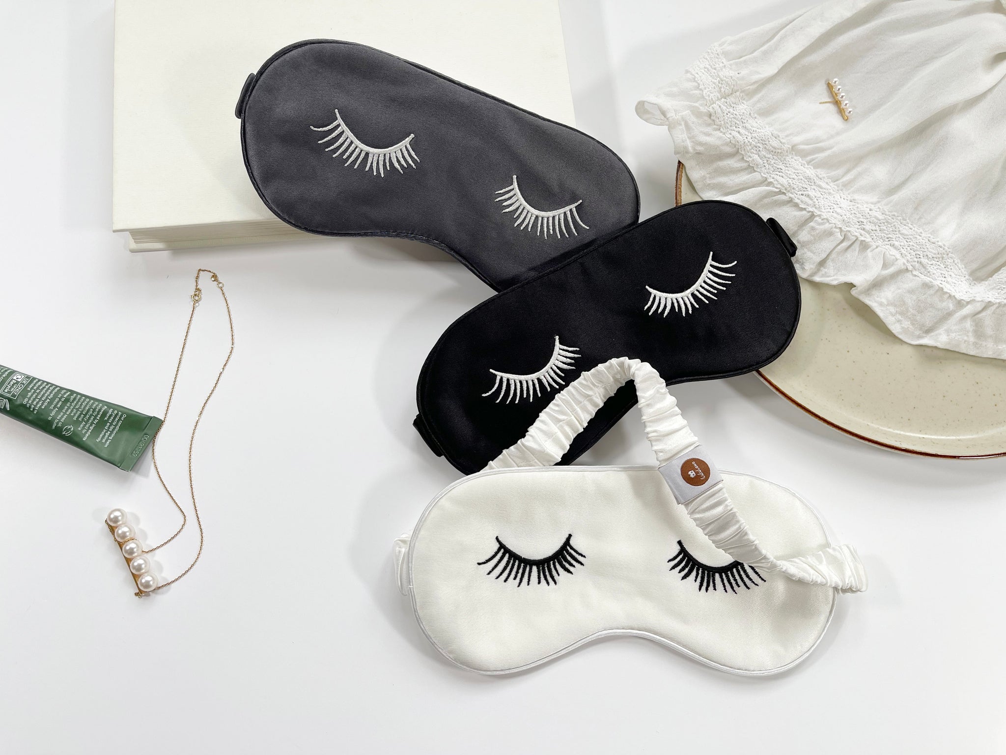 three silk eye masks featuring eyelashes embroidery and elastic strap in grey, black and white colors 