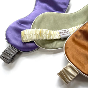 three professionally designed and handcrafted silk eye masks with elastic straps. One is purple with grey strap, one is pea green with creamy white strap and another one is burnt orange with champagne strap